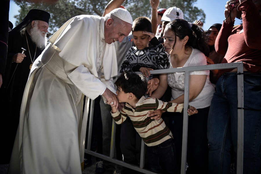 In questa immagine ricevuta dall’ufficio stampa del Primo Ministro, un bambino bacia la mano di Papa Francesco durante la sua visita al centro di detenzione per migranti e rifugiati di Moria vicino a Mitilene, sull’isola di Lesbo, il 16 aprile 2016. Dichiarando “siamo tutti migranti”, Papa Francesco ha portato un messaggio di speranza a migliaia di persone che rischiano l’espulsione dalla Grecia mentre criticava la comunità mondiale per non essere riuscita a porre fine alle guerre che alimentano la crisi. In una visita emozionante che ha visto persone inginocchiate in lacrime ai piedi del papa, il pontefice ha detto agli esuli che “non sono soli” e ha chiesto al mondo di mostrare “comune umanità” a seguito dell’irrigidimento della posizione dell’UE sui migranti. (Photo by ANDREA BONETTI / HANDOUT / AFP) (AFP)