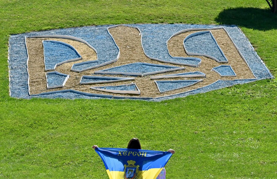 Una giovane donna tiene la bandiera ucraina davanti all'emblema nazionale ucraino in un prato di Kiev il 23 agosto 2025, un giorno prima della Giornata dell'indipendenza dell'Ucraina, in mezzo all'invasione russa in Ucraina. (Sergei Supinsky / AFP)