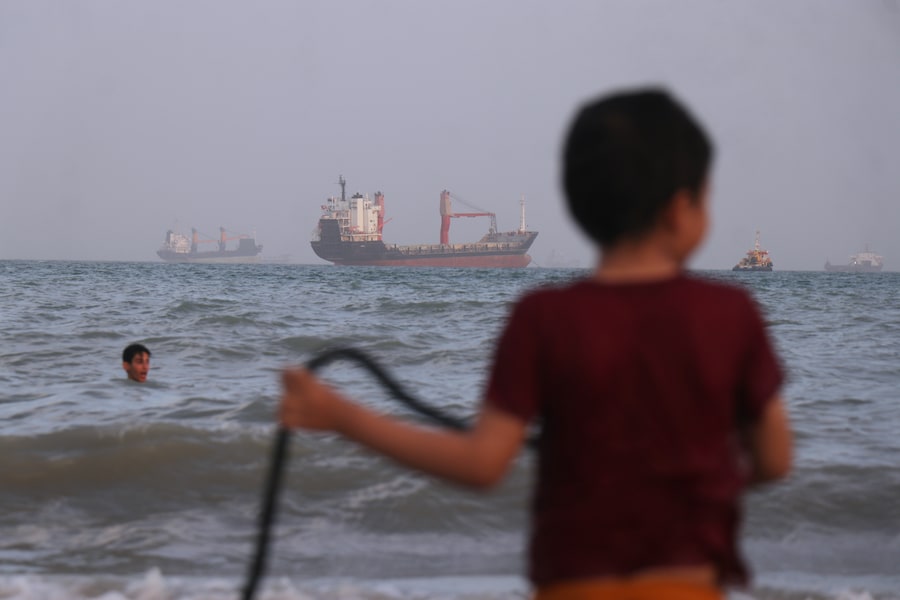 I bambini giocano nell'acqua lungo la riva mentre una flotta composta da navi portarinfuse, navi da carico e imbarcazioni di servizio è ormeggiata al largo dello Stretto di Hormuz. (APN)
