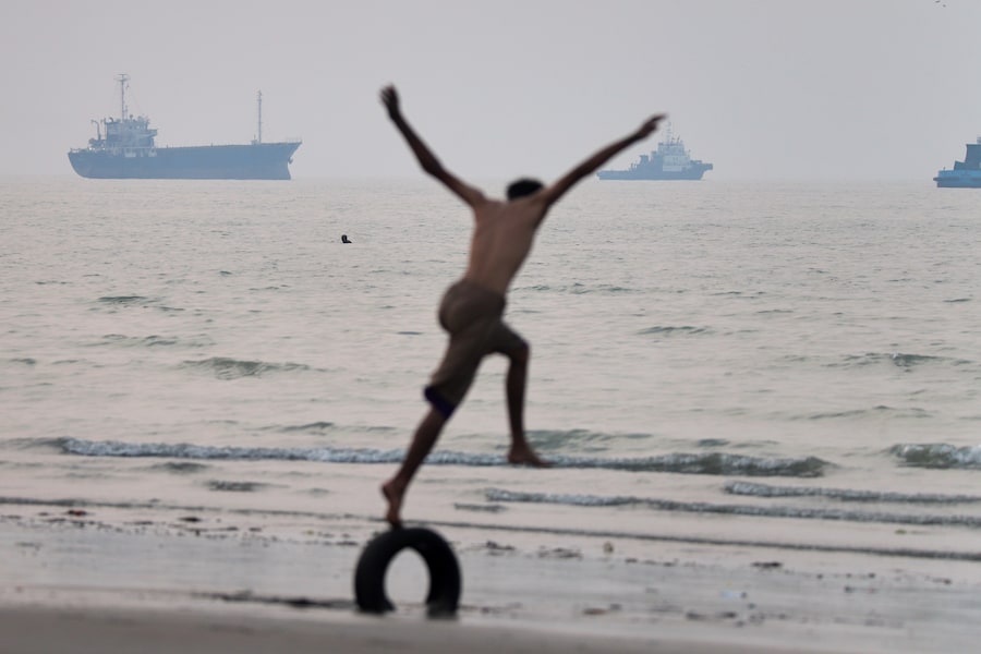 Un ragazzo sta in equilibrio su uno pneumatico in riva al mare, mentre si intravedono delle navi nello Stretto di Ormuz al largo di Bandar Abbas, in Iran, martedì 30 giugno 2026. (APN)