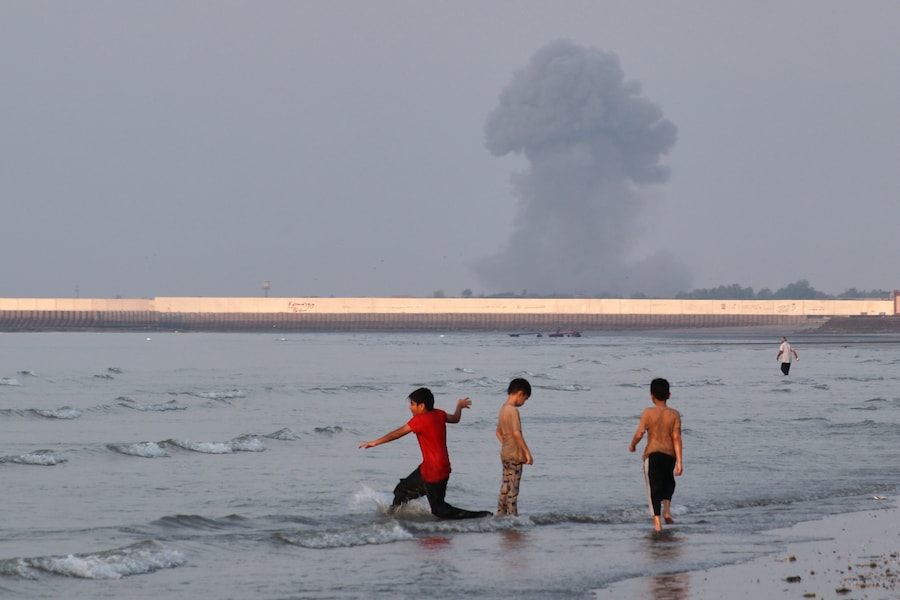 Tre ragazzi giocano nelle acque poco profonde dello Stretto di Hormuz, mentre sullo sfondo si alza una colonna di fumo causata da un’esplosione al largo di Bandar Abbas, in Iran. (APN)