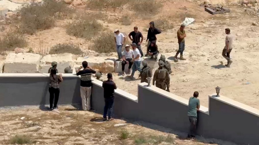 I palestinesi usano i loro telefoni mentre coloni israeliani e militari si trovano davanti a un'abitazione palestinese a Qusra, nella Cisgiordania occupata da Israele. (via REUTERS)