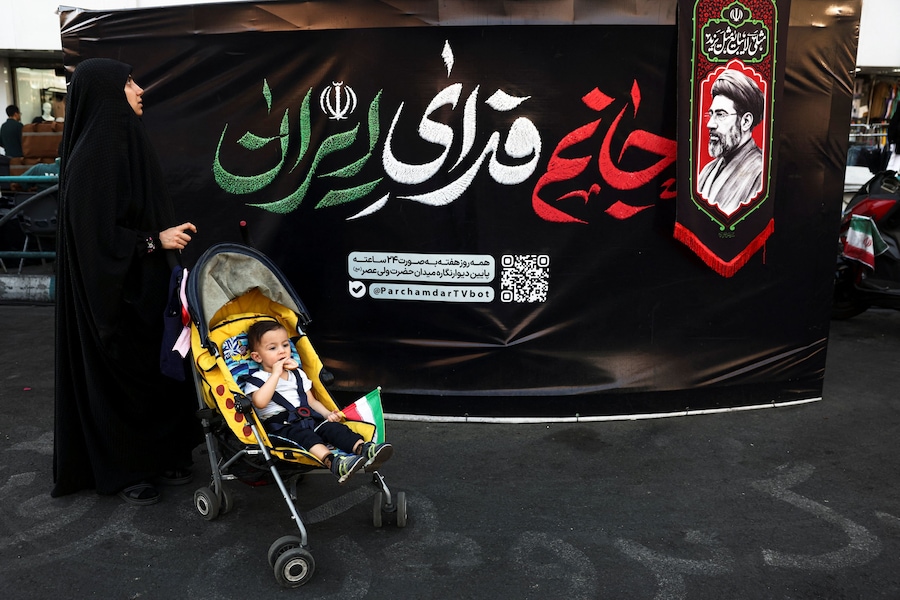 Una donna con il proprio bambino in un passeggino è in piedi accanto a una foto della Guida Suprema dell’Iran Mojtaba Khamenei, in una strada di Teheran. (Majid Asgaripour/WANA (West Asia News Agency) via REUTERSS)