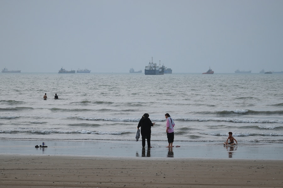 Alcune persone trascorrono del tempo in spiaggia mentre navi commerciali sono ancorate nello Stretto di Hormuz al largo di Bandar Abbas, in Iran. (APN)