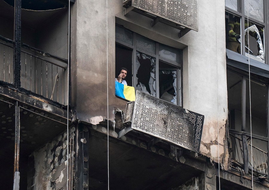 Un uomo espone la bandiera nazionale dalla finestra del suo appartamento danneggiato sul luogo di un attacco con droni russi contro un grattacielo residenziale a Kiev (EPA)