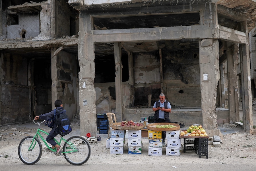 Un ragazzo in bicicletta guarda verso un venditore di frutta che sta usando il cellulare in una bancarella lungo la strada, davanti a un edificio danneggiato durante la guerra civile a Homs, in Siria, giovedì 1 ottobre 2026. (Foto AP/Ghaith Alsayed) (APN)