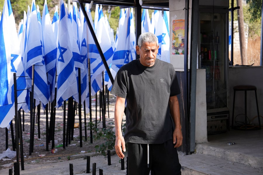 Il proprietario palestinese di una caffetteria, Mueyyid Hussein, si trova davanti al suo locale mentre nelle vicinanze sventolano bandiere israeliane issate dai coloni, che secondo lui hanno ostruito l’ingresso e la visibilità del locale, ad Al-Lubban al-Sharqiya, vicino a Nablus, nella Cisgiordania occupata da Israele, il 9 ottobre 2026. REUTERS/Ali Sawafta (REUTERS)