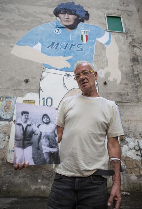 Antonio «Bostìk», barista, in posa davanti al murales storico di Maradona del 1990 a largo degli Artisti nei Quartieri Spagnoli, mostrando la foto in cui, da giovane ultras, avvicinò il capitano a bordocampo al termine di una partita. Napoli, agosto 2020. © Paolo Vezzoli