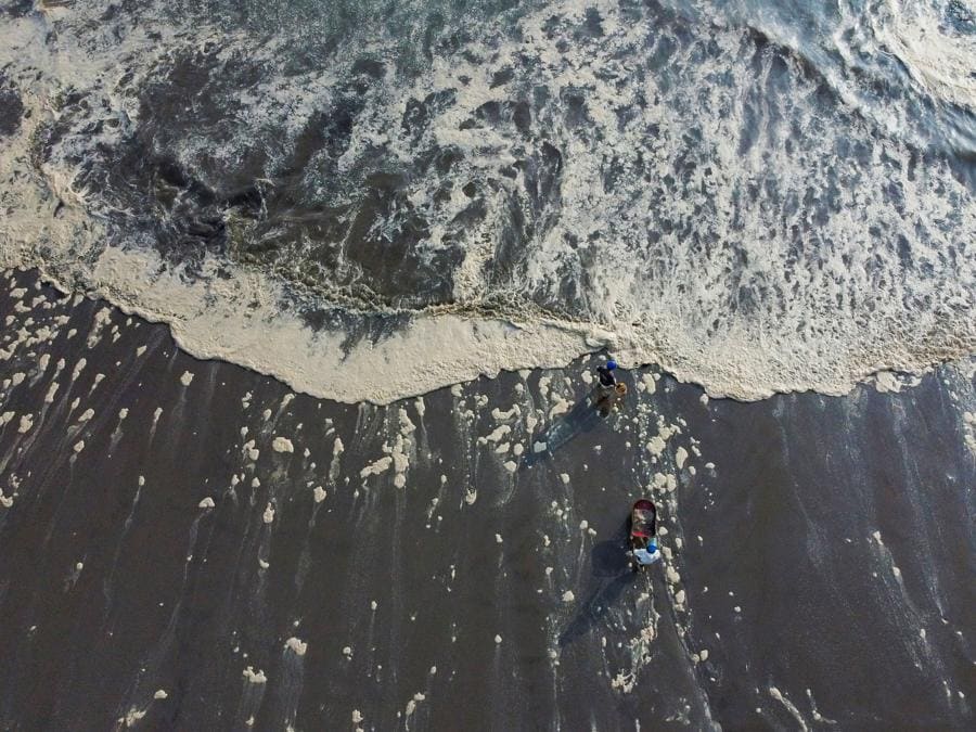 Lavoratori camminano attraverso la schiuma su una costa colpita da una fuoriuscita di petrolio causata da onde anomale, innescata da una massiccia eruzione vulcanica sottomarina a Tonga, al largo della costa di Lima, a Ventanilla, Perù, 19 gennaio 2022. Foto scattata con un drone.(REUTERS/Angela Ponce)