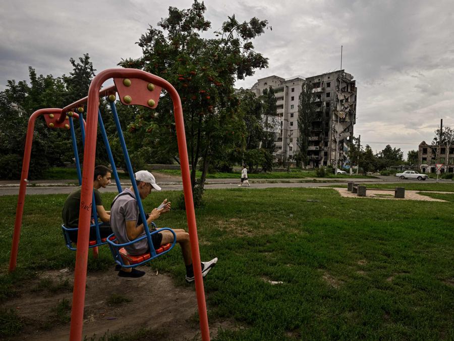 Due giovani uomini sulle altalene in un parco giochi di fronte a un edificio residenziale distrutto nella città di Borodyanka. (Photo by Genya SAVILOV / AFP)