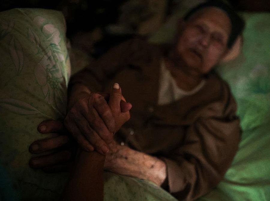 Natalia Ataiantz, 42 anni, tiene le mani di sua nonna Mariia Tkachenko, 96 anni, sdraiata sul suo letto all’interno della loro casa vicino a Bakhmut.  REUTERS/Nacho Doce