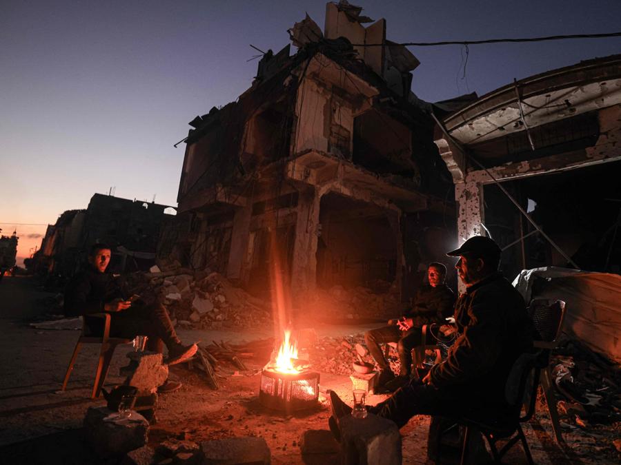 Le persone si siedono attorno a un fuoco da campo in mezzo agli edifici distrutti nel distretto di Khezaa, alla periferia della città di Khan Yunis, nel sud della Striscia di Gaza. (Photo by MAHMUD HAMS / AFP)