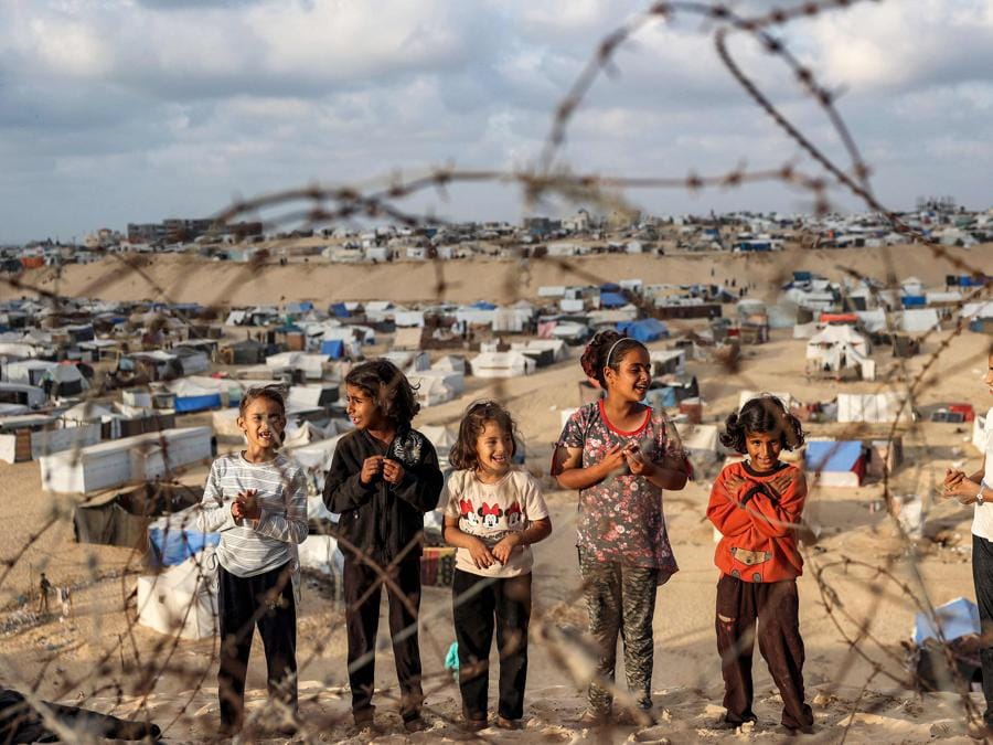 Bambini in piedi dietro il filo spinato lungo un pendio vicino a un campo che ospita palestinesi sfollati a Rafah, nel sud della Striscia di Gaza. (Photo by AFP)