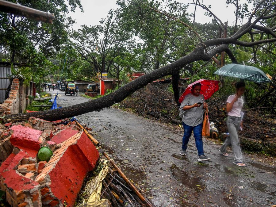 Persone con ombrelli in mano passano davanti a una casa danneggiata e a un albero sradicato durante la pioggia a Guwahati, in seguito all’approdo del ciclone Remal in India. Il ciclone Remal, che si è abbattuto sulle pianure del Bangladesh e sulla vicina India la sera del 26 maggio con violente tempeste e onde che si infrangono, ha provocato la morte di almeno 38 persone, distrutto migliaia di case, distrutto dighe marine e allagato città nei due paesi. (Photo by Biju BORO / AFP)