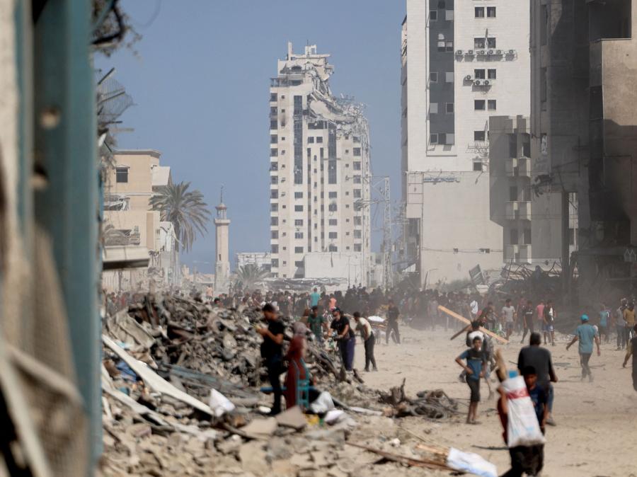 La gente passa davanti alle macerie, con la vista della Torre Mushtaha danneggiata sullo sfondo, in seguito al ritiro delle truppe israeliane dal quartiere Al-Sinaa della città occidentale di Gaza. (Photo by Omar AL-QATTAA / AFP)