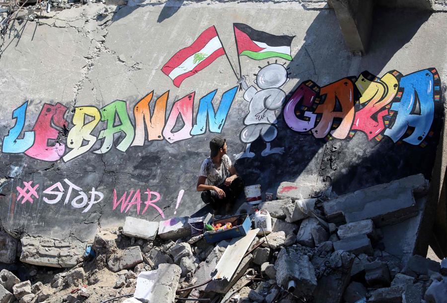 Un giovane palestinese siede accanto a un murale che ha dipinto sul tetto di una casa distrutta a Khan Yunis, nel sud della Striscia di Gaza, per mostrare la sua solidarietà con il popolo di Gaza e  Libano.  (AFP)