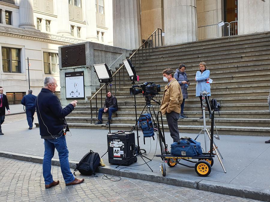 La troupe di una tv finanziaria davanti a Wall Street: un reporter attende il collegamento per raccontare la giornata di Borsa. Dietro la telecamera l'operatore indossa la mascherina.